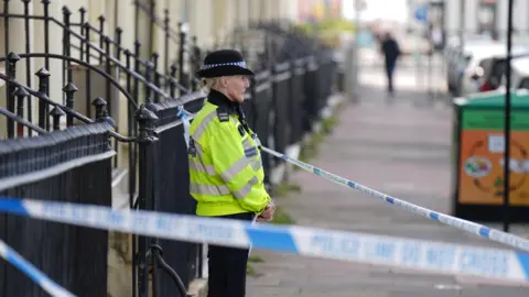 Eddie Mitchell A police officer outside of a building behind police tape 