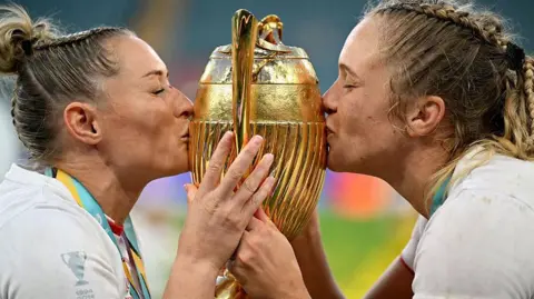 Dan Mullan/Getty Images Two female rugby players, wearing white England kits and with medals around their necks, are either side of a gold trophy kissing it.