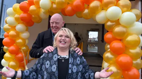 A smiling Steve Herron, who has a bald head and is wearing a black woollen coat, poses with his hands on the shoulders of Sue Herron, who has blonde hair and and has her arms outstretched. In the background is an archway made of orange, golden and yellow balloons. 