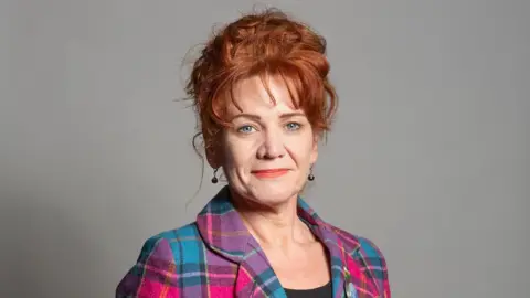 UK Parliament A headshot of Sarah Atherton. She has ginger curly hair which is tied back, a fringe, and blue eyes. She is wearing coral lipstick, round drop earrings and a pink and blue plaid jacket. She is standing in front of a grey background. 