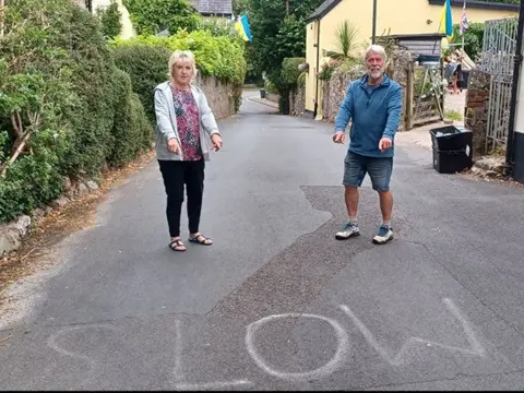 Marilyn and Norman Roberts stand on a narrow village road beside the word "SLOW" hand written on the tarmac, gesturing toward it. The street is lined with hedges and stone walls, with houses visible in the background.