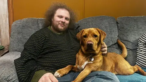 Rhys and Eddie sit side by side on a grey sofa, and both are looking at the camera. Eddie is a golden-coloured labrador and is sitting with his front legs over Rhys's lap.