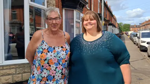 LDRS Two ladies, one with a flowery patterned dress, short hair and glasses and the other with a blue jumper and shoulder length hair. They are both smiling. They are stood on a residential street with parked cars.