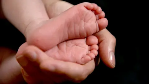 A mother's hand holding the feet of an infant against a black backdrop.
