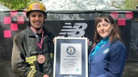 A man is dressed in firefighter uniform with a black jacket and yellow helmet, wearing a marathon medal around his neck is being presented with a framed certificated by a woman on his left dressed in a blue blazer. She has brown shoulder-length hair