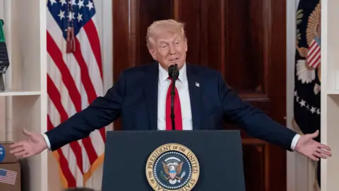 Donald Trump pictured speaking wearing a blue suit and red tie at an event at a podium marked with a circular White House logo. Behind him, there are objects as part of an event highlighting business investment in the US 