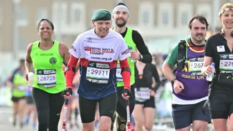 ROYAL NAVY & ROYAL MARINES CHARITY Chris Terrill taking part in the Brighton Marathon on crutches. He is wearing a brightly coloured t-shirt and a green beret.