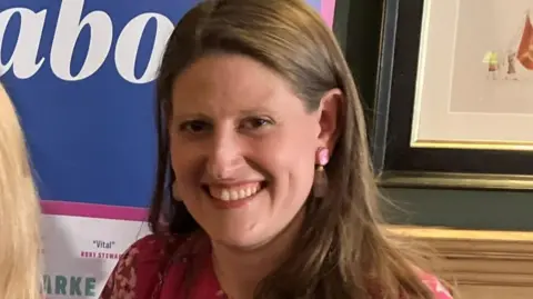 Theo Clarke stands in front of a banner adverting her book. She has shoulder-length dark hair and is wearing a pink dress and has pink earrings.