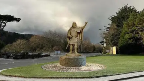 A mock-up of the statue of King Orry in place at St John's. The golden artwork is of a man wearing draping clothes, with a large beard and a hat, who holds a sword and raises his left arm. It stands on a plinth on grass near to a road.