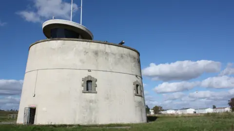 Jaywick Martello Tower Jaywick Martello Tower, which is a circular fort made of white stone. It is set in a field and has been pictured on a bright day.