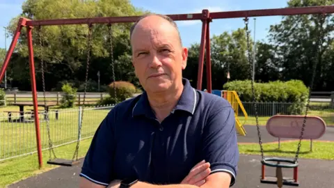 BBC / Jasmine Lowe The photo shows a man looking at the camera. His arms are folded across his chest. He's wearing a blue polo shirt. He's standing in a play area in front of some swings. There is also a slide, lots of grass, and a picnic bench behind him. 