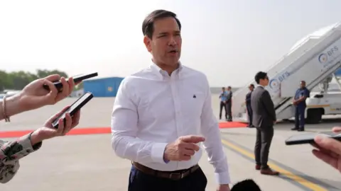 Marco Rubio, wearing a white shirt and dark trousers, on the tarmac of an airport, point with his finger as he speaks to journalists - not in shot, just a couple of hands with phones recording him. Other people can be seen in the background, near the stairs of an airplane.