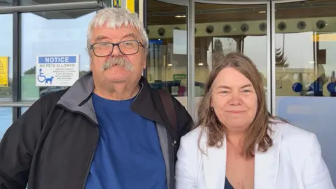 Martin and Ros Hughes pictured standing side by side outside an airport entrance. Martin on the left is wearing a navy T-shirt and a black jacket. He has short grey hair, a moustache and is wearing glasses. Ros is wearing a white linen blazer and has short brunette hair.