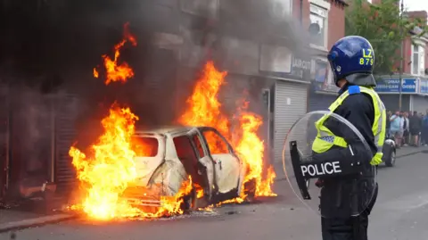 PA Media A police officer wearing a helmet and shield looks at a white car on fire outside a shop.