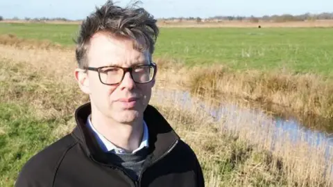 John Fairhall/BBC Eliot Lyne is standing outside by a ditch in a field. He is standing on the left of the frame and is looking directly at the camera. He is wearing glasses and is wearing a black sweatshirt, shirt and grey jumper. 