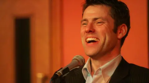 Stuart Steele/Civic Comedy A man with dark hair in an open necked shirt under a striped suit jacket is in front of a microphone. He is laughing