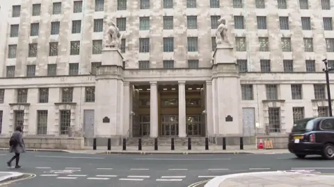 The Ministry of Defence main building on Whitehall in London, with its stone statues flanking the columned entrance and a black cab passing
