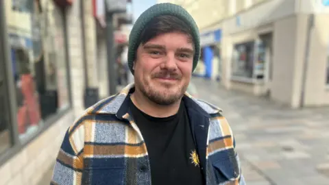 Elliot Kinrade in  Strand Street smiling at the camera. He is wearing a teal-coloured hat, a blue and yellow checked shirt and a black t-shirt.