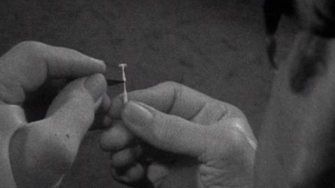 Black and white close up image of the sculptor's hands.  Part of the plastic support on his wrists are visible.  Within his fingers, a partially carved matchstick is visible in his right hand and part of a razor in his left hand is working on the match.  The sculptor's left ear, sideburn and spectacles are visible in the right hand foreground.