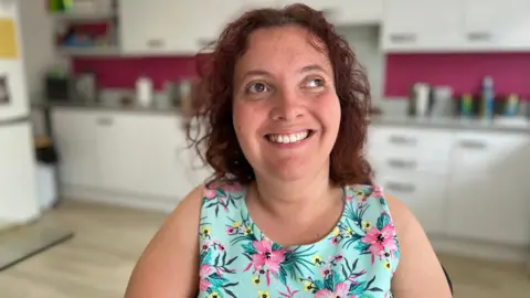 A person sits in a kitchen with white cabinets and a pink wall. They are wearing a dress with a floral pattern of pink flowers and green leaves. Various kitchen items are visible on the counter.