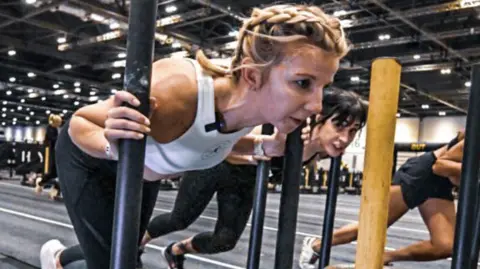 Hyrox Tor during a Hyrox race, pushing some equipment, in a room, like a gym, with two people also exercising. She has a concentrated face. Her blonde hair is tied back in braids. 