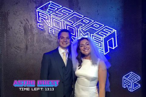 Escape Rooms Cardiff Harmony and Seán smiling at the Escape Room. Behind them is a backdrop with Escape Rooms lit up in large blue writing. On the bottom left corner, there is writing showing how long they have left on the Escape Room.