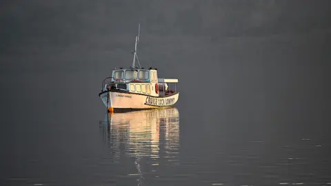 Victor Tregubov A small white boat sits still on calm, reflective water in soft evening light, with faint hills forming a dark backdrop.