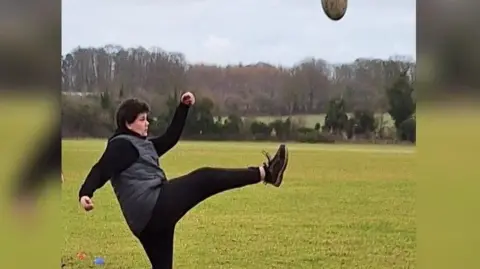 Oakland School Student Isaish in black trousers, top and trainers kicking a rugby ball on a field.