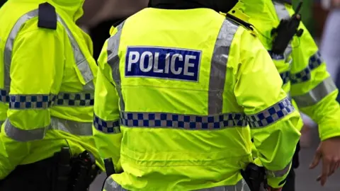 The back of three police officers wearing yellow hi-vis jackets which say police on the back.