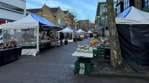 The market on High Street in Staines. Several different stalls with white and blue covers, with a fruit and vegetable stall very noticeable in the foreground.