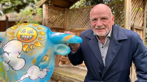 John Harris wearing a blue coat and patterned shirt, crouching down next to a ceramic bull, which he bought at auction for Birmingham Hospice