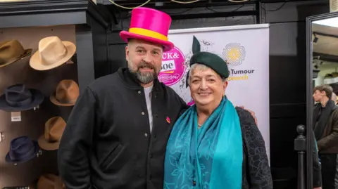 Brain Tumour Research Alfie Boe and Anne McEntee. He is wearing a pink top hat and she is wearing a blue scarf and green hat