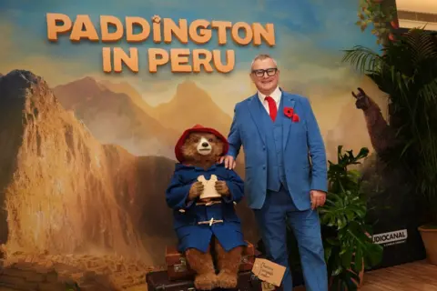 Hugh Bonneville poses for a photo with Paddington Bear in front of a large poster advertising the new film Paddington in Peru. He wears a light blue suit with matching waistcoat and trousers, a bright red tie and a red poppy on the lapel. He has thick dark glasses and is smiling. paddington is holding a sandwich with a chunk bitten out, and wears his blue duffel coat and red hat. The poster shows mountains and jungle in the background.