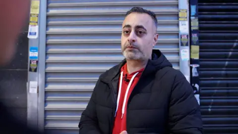 Kuldeep Maan,  wearing a black padded jacket over a red hoodie stands in front of a closed metal shop shutter, with various small stickers and notices posted on the frame beside it