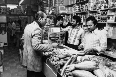 Getty Images Black and white photo of shopkeeper Komor Uddin serving a customer at his grocer's shop, the Taj Stores in Brick Lane