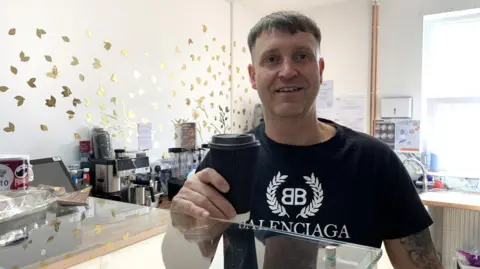 BBC/Lucy Ashton A man with short, dark cropped hair is wearing a black t-shirt with a white slogan. He is smiling and holding a disposable coffee cup with a lid over a coffee shop counter