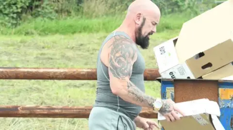 Nottinghamshire Police A man with a beard and tattoos holds a box next to a skip
