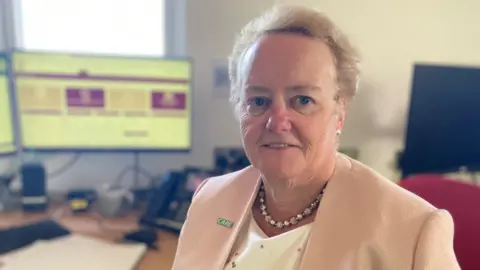 A woman with short ginger-blond hair sits in an office in front of computer monitors. She is wearing a pink jacket, white top and chunky silver necklace.