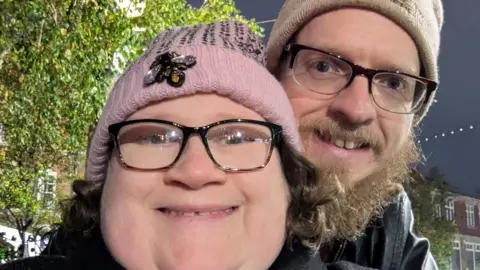 A woman with black rimmed glasses and a pink bobble hat smiles at the camera. Behind her a man with a beige bobble hat cuddles up to her. He has black rimmed glasses too and a dark beard.