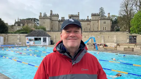 A middle-aged man wearing a grey and red coat and a dark cap with a white emblem on smiles as he stands in front of a pool with lanes. There is a large, castle-like stately home on the other side of the wall.