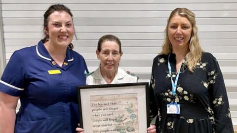 Three women stand together, Robyn Dalton on the left is wearing a blue midwife's uniform, Carol Goddard in the centre is wearing a white healthcare assistant's uniform and holding a framed quotation. Jo Hadley is on the rightand wearing a blue dress with yellow flowers and an NHS lanyard
