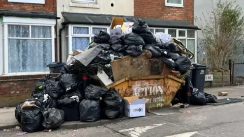 A skip overflowing with bin bags. The street has cobbling in parts and the yellow skip has "action" stencilled on the side. A cardboard box stands in front while dozens of bin bags are piled over the skip. Behind the skip are several homes with net curtains in their front windows.