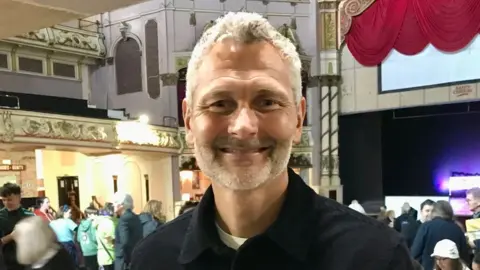 LDRS Si Bellamy standing smiling in a hall in Morecambe with people in the background milling about