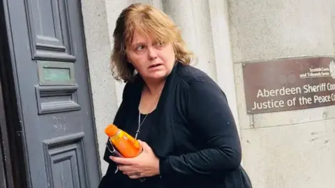 Shirley Freeman, a woman dressed in black and holding an orange bottle of fizzy juice, arriving at Aberdeen Sheriff Court.