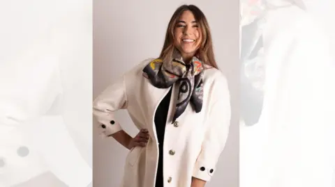 A woman with long, dark hair smiles and places a hand on her hip as she poses in a cream-coloured designer coat and black patterned silk scarf.