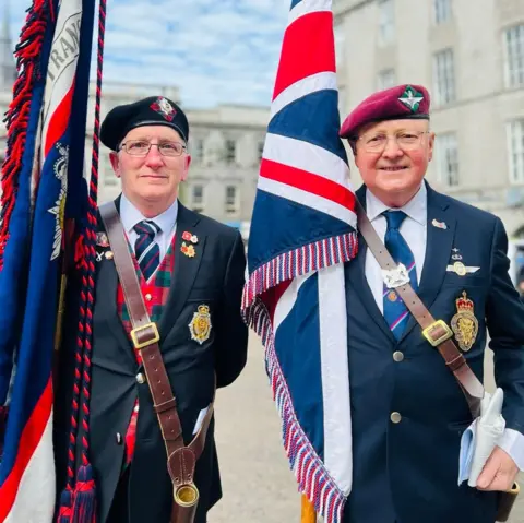 Two men face the camera wearing suits and military regalia carrying large flags