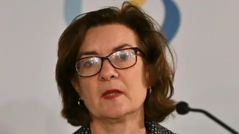 Close up image of Eluned Morgan talking at a government event. She is talking whilst looking out at the audience. She has brown short hair and is wearing glasses. 
