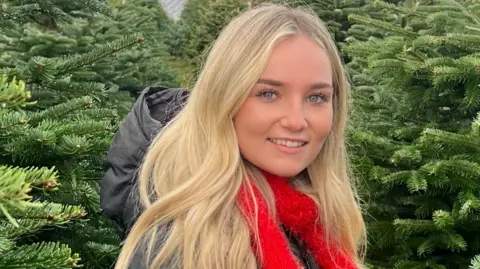 Family photo Emily is standing in a grove of Christmas trees. she is wearing a grey parka and has a red scarf tied at her neck. She has long wavy blonde hair, blue eyes and is wearing light makeup. 