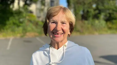 A lady with brown hair looking at the camera. She's wearing a white hoodie with a drawstring.