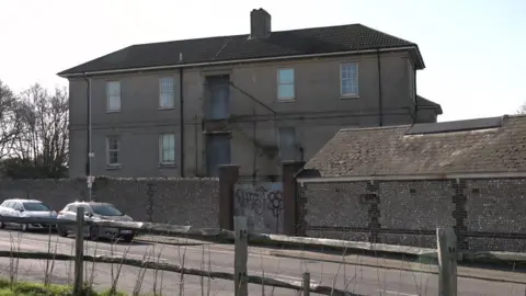 A large grey building. There are a couple of cars parked on the street. 
